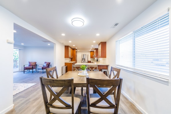 A modern dining area with a wooden table and chairs, a kitchen in the background, and a large window with blinds. - Beautiful like new home with a large hot tub in a quiet neighborhood on Acreage. (Oregon City)