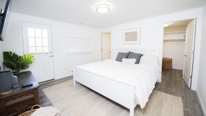 Ein Schlafzimmer mit einem weißen Bett, einem Fernseher auf einem Holztisch, einer Pflanze und einer Tür zu einem anderen Raum.