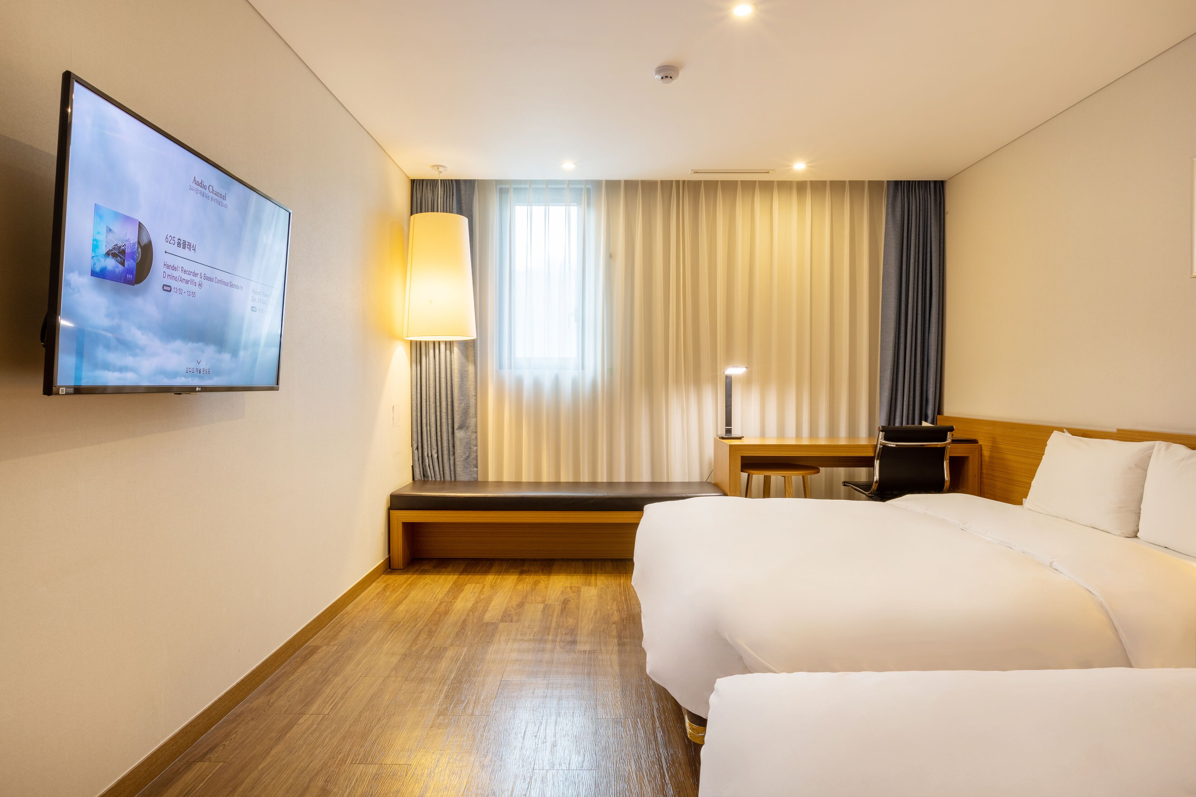 Habitación de hotel con cama, escritorio, lámpara, televisor en la pared y ventana con cortinas.