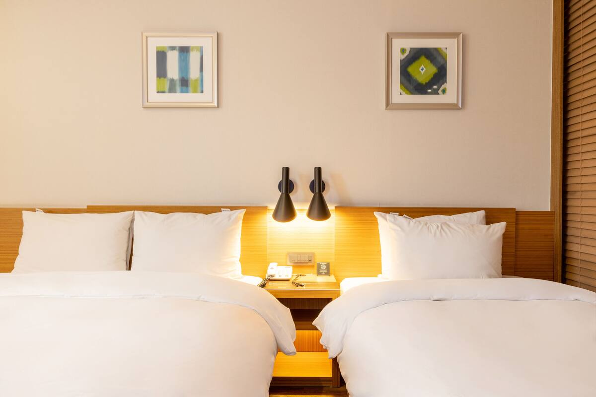 A double bed with white bedding, two bedside tables, a wall-mounted lamp, and two framed artworks on the wall.