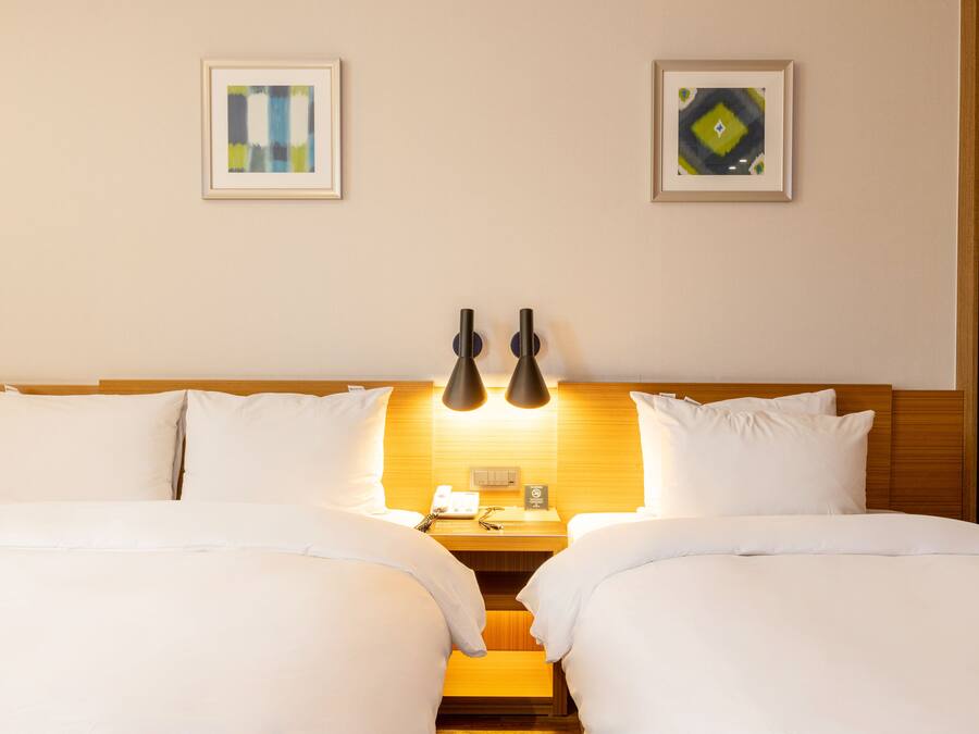 Una cama doble con ropa de cama blanca, dos mesitas de noche, una lámpara de pared y dos cuadros enmarcados.