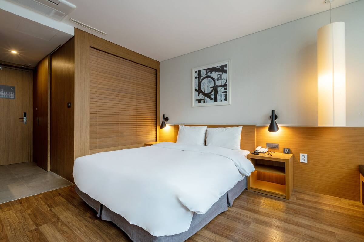 A modern hotel room with a large bed, wooden headboard, a wall-mounted artwork, and a bedside table with a lamp.