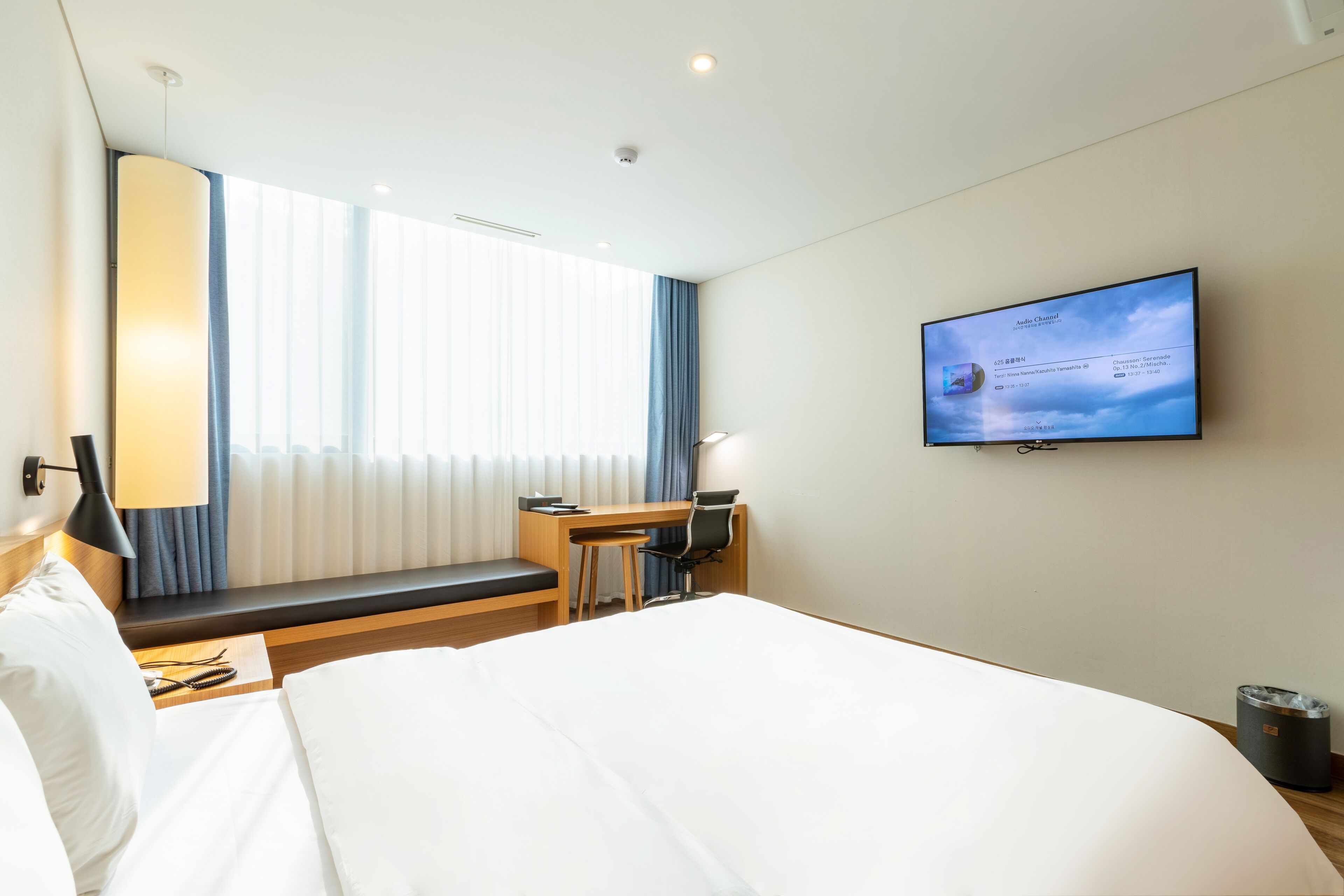 A hotel room with a large bed, a desk, a chair, a television mounted on the wall, and a window with curtains.