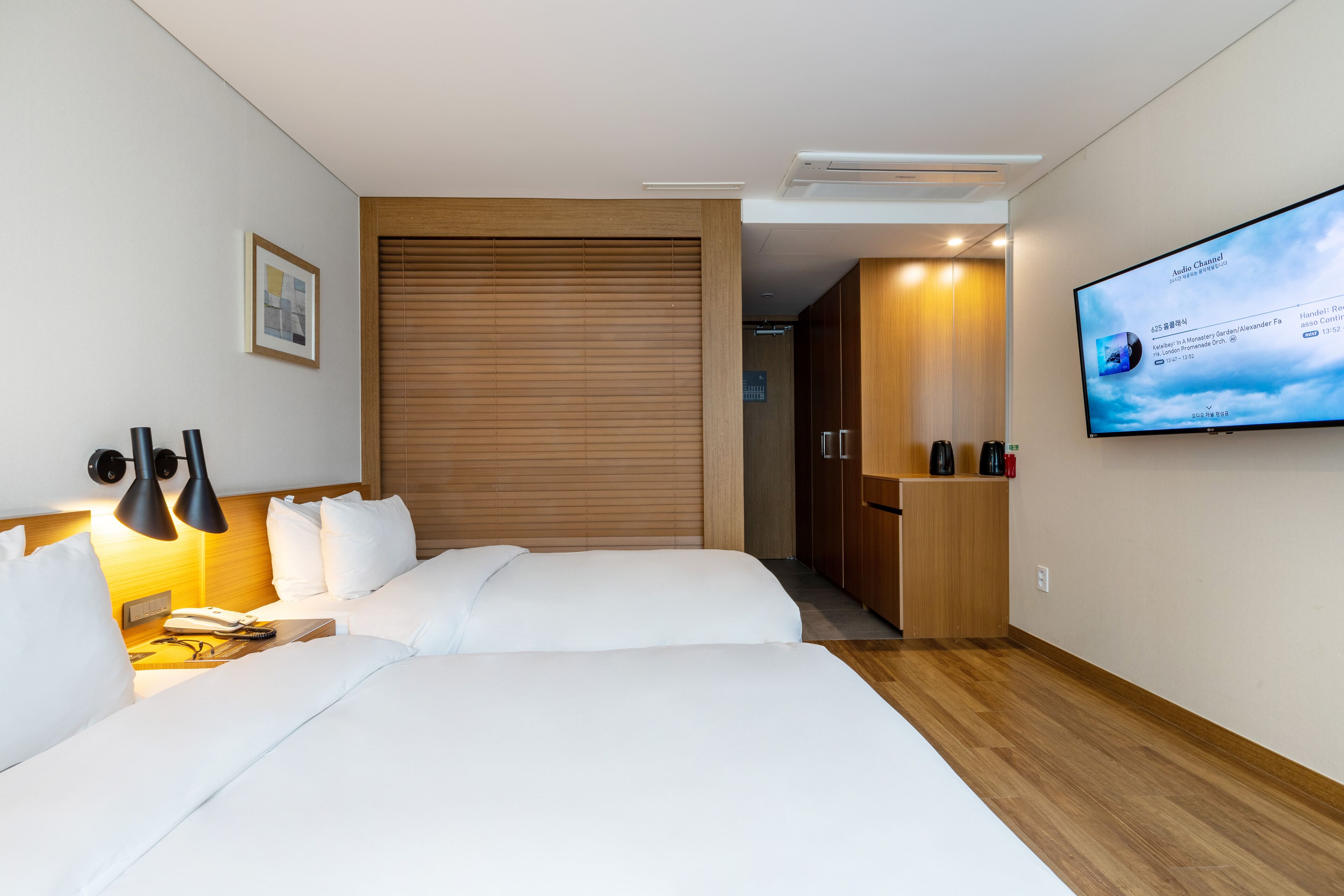 Una habitació d'hotel amb dos llits, un televisor fixat a la paret, un armari de fusta i una taula de nit amb làmpades.