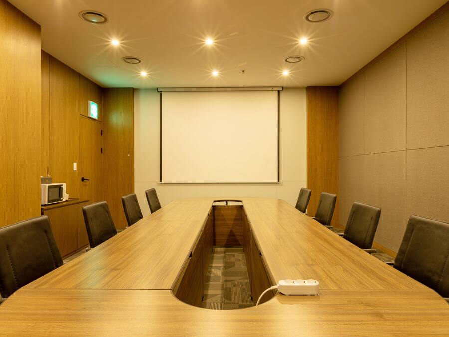 Un salón de conferencias con una larga mesa de madera, ocho sillas, una pantalla de proyector y un microondas sobre un mueble.