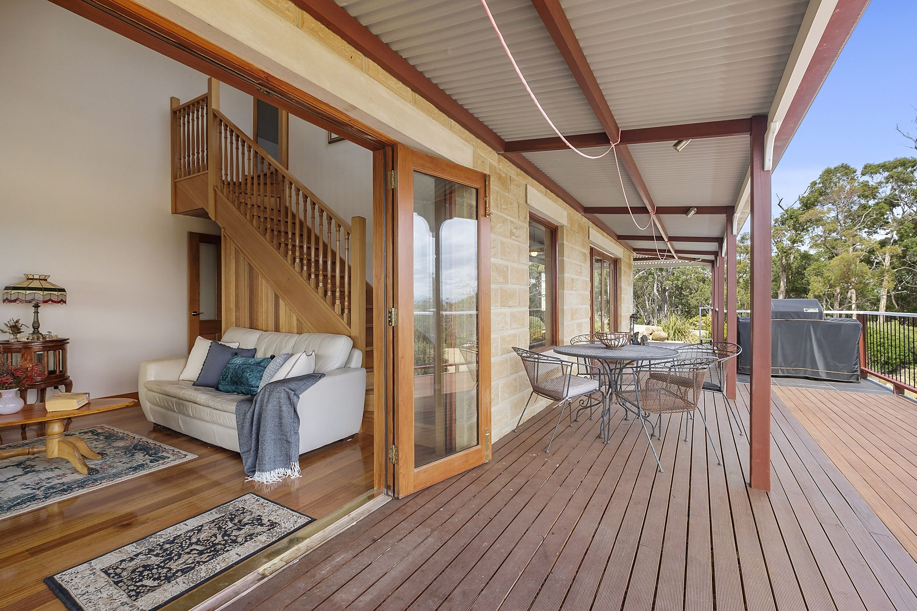 A covered outdoor area with a wooden deck, a sofa with cushions, a dining table with chairs, and a staircase leading to an upper level.
