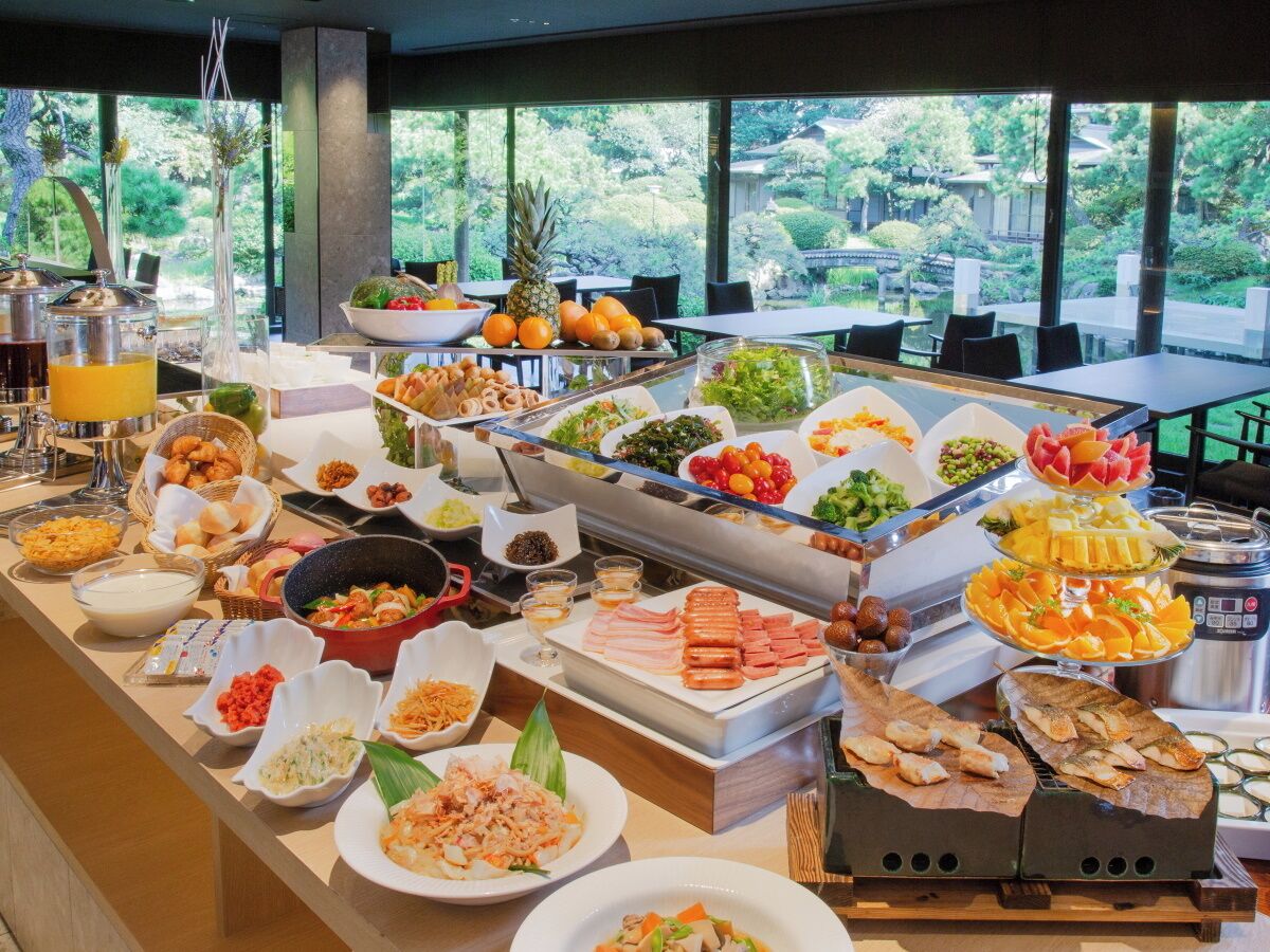 A buffet with various food items and a view of a garden through large windows.