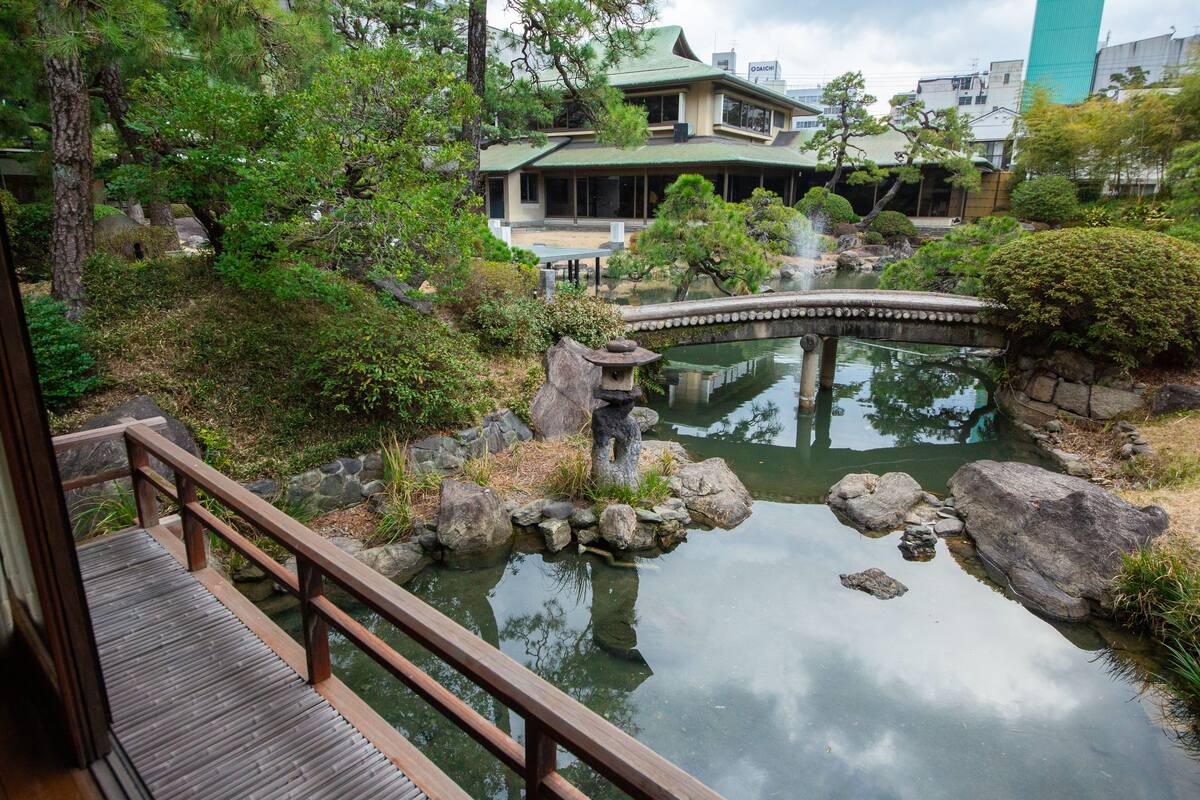 A traditional Japanese garden with a wooden bridge, a pond, and a building with a green roof.