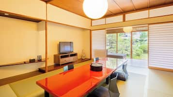 Una habitación tradicional japonesa con suelo de tatami, una mesa baja con superficie roja, un televisor sobre un soporte de madera y vistas a la vegetación exterior.