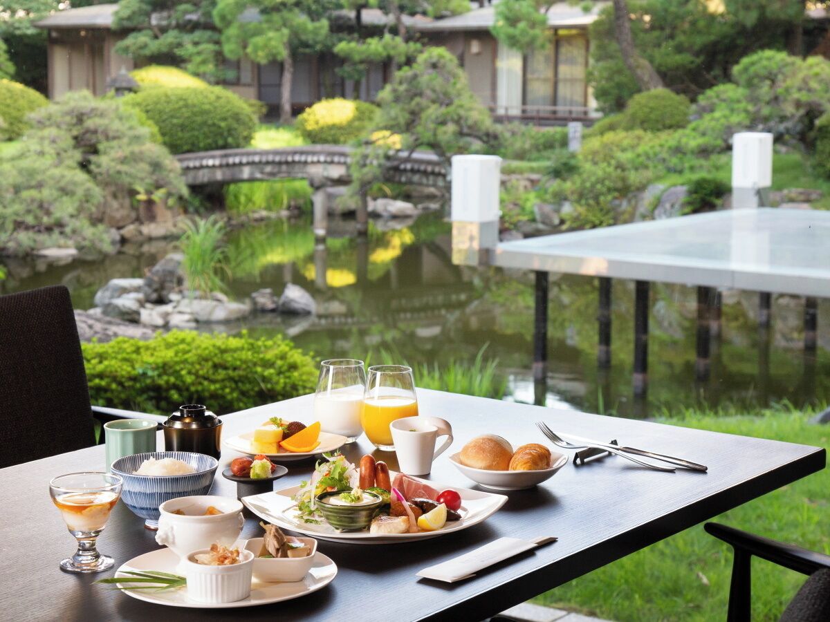 A dining table with a meal set, including plates, bowls, and glasses, overlooking a garden with a pond and traditional architecture.