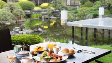 A dining table with a meal set, including plates, bowls, and glasses, overlooking a garden with a pond and traditional architecture.