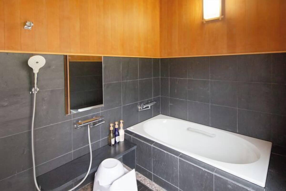A modern bathroom with a large bathtub, dark tiles, wooden paneling, a handheld showerhead, and a small mirror.