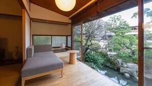 A modern room with a wooden floor, a bed, a small round table, and a view of a garden through large windows. - Suisui Garden Ryokan (Kitakyushu)