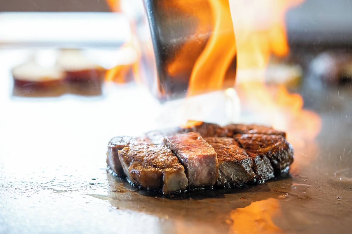 A steak being grilled on a flat surface with flames visible in the background.