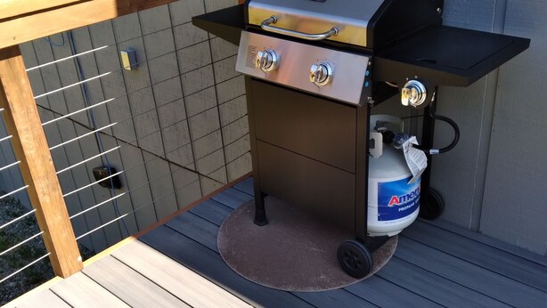 Una parrilla para barbacoa con dos quemadores, un tanque de propano y una terraza de madera.