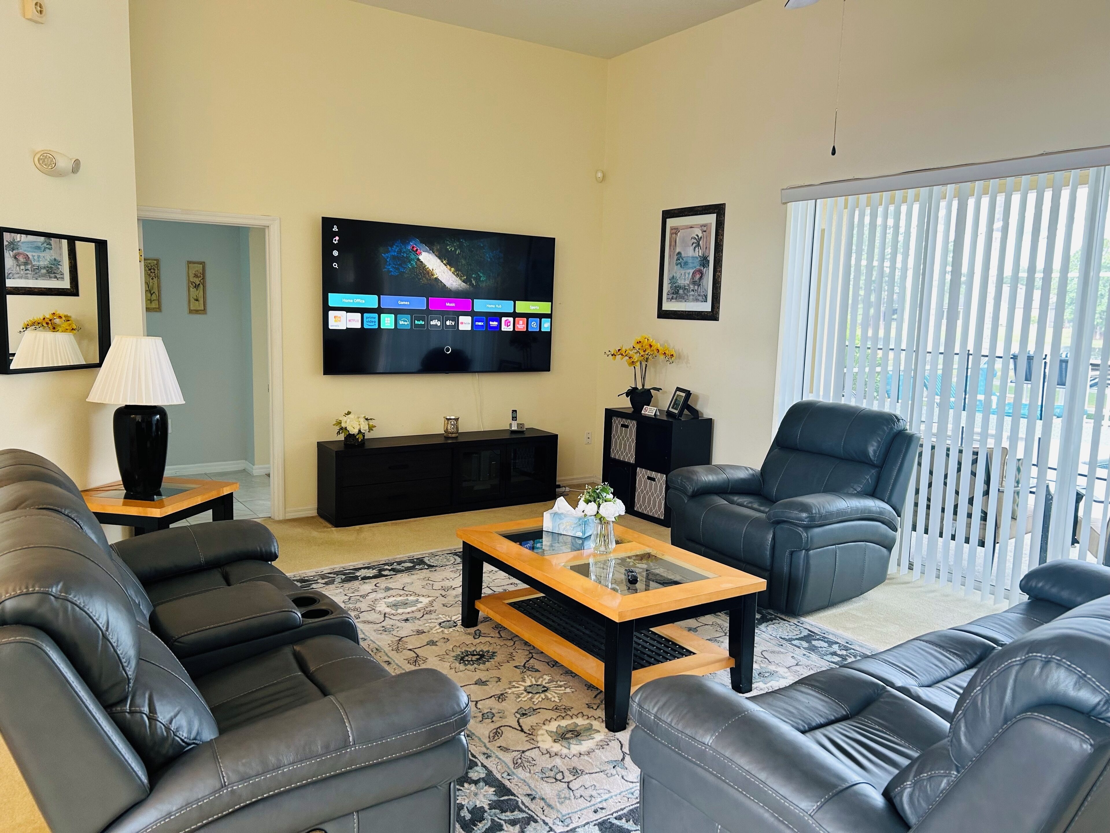 A living room with a flat-screen TV, black leather sofas, a coffee table, and a large window with blinds.
