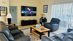 Una sala de estar con televisor de pantalla plana, sofás de cuero negro, una mesita de centro y un ventanal con persianas.
