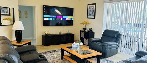 A living room with a flat-screen TV, black leather sofas, a coffee table, and a large window with blinds.