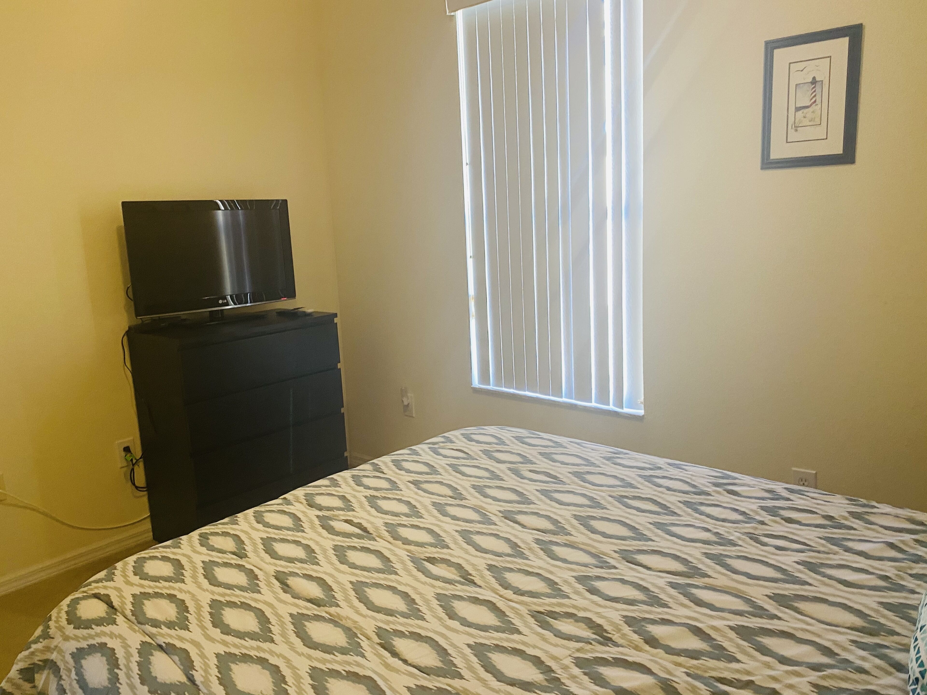 Un dormitorio con cama, cómoda, televisor encima de la cómoda, ventana con persianas y un cuadro en la pared.