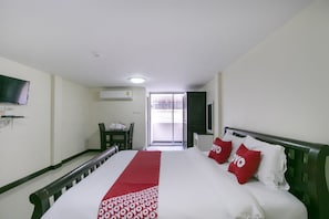 A hotel room with a bed, a TV mounted on the wall, a desk with a chair, and a window with a view of the outside.