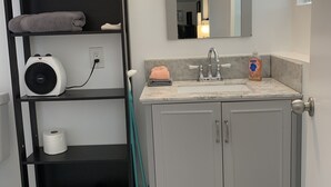 A bathroom with a grey vanity, a towel rack with folded towels, a mirror, a towel dispenser, and a fan on a shelf. - Shabby Chic Studio in LA Hills (Alhambra)