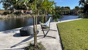 A concrete pathway with a hammock and a black bean bag chair, leading to a body of water with a fence and houses in the background.