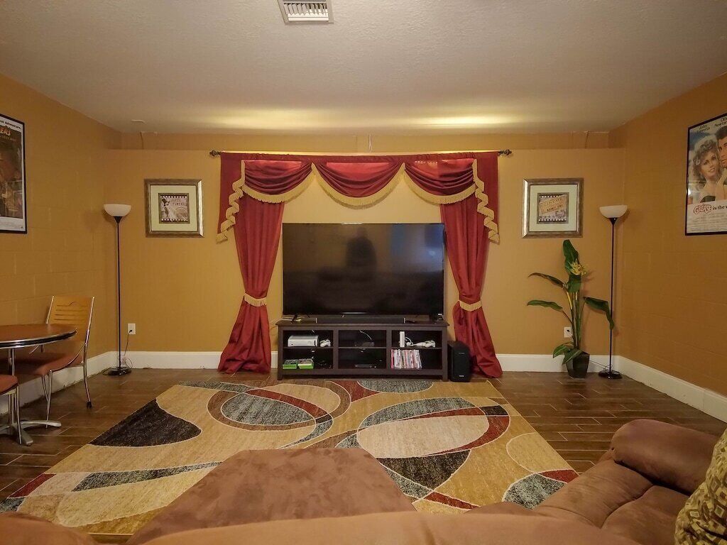 A living room with a flat-screen TV, red curtains, a patterned rug, and a brown sofa.