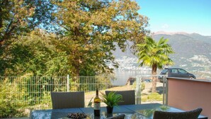 Una mesa con cubierta de vidrio, sillas de mimbre, una botella y un cuenco con nueces en un patio con vistas a montañas y un coche al fondo.