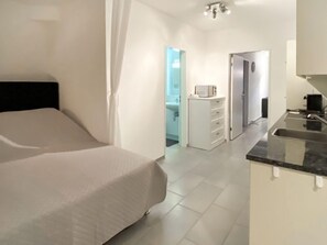 Une chambre d’hôtel moderne équipée d’un lit, d’une kitchenette et d’une salle de bain, le tout visible à travers une porte ouverte.
