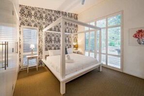 A bedroom with a four-poster bed, a bedside table with a lamp, a large window with blinds, and a patterned wallpaper. - Tonic—Family Bushland Retreat near Yallingup Beach (Yallingup)