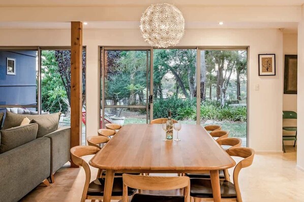 A dining area with a wooden table and chairs, a chandelier, and a view of greenery through large sliding doors.