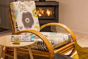 A wicker chair with a cushion, a small round table with a mug and a magazine, and a wood-burning stove in the background. - Tonic—Family Bushland Retreat near Yallingup Beach (Yallingup)