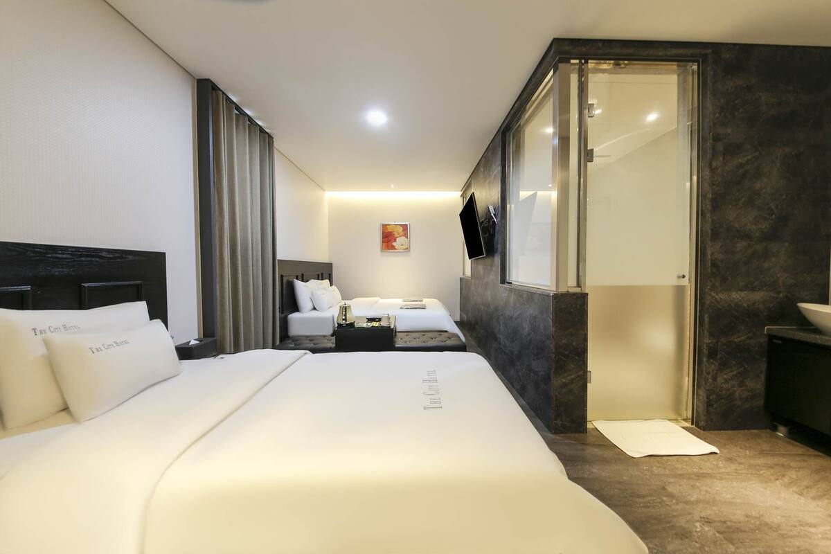 Habitación de hotel con una cama grande, una cama más pequeña, un escritorio, una silla, un televisor y un baño con espejo y lavamanos.