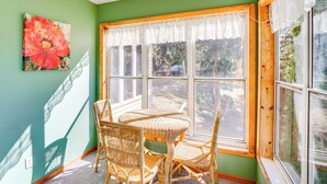 Ein Zimmer mit grüner Wand, einem großen Fenster mit Spitzenvorhängen, Korbmöbeln und einem Gemälde einer roten Blume.