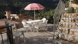 A terrace with a table and chairs, a red and white striped umbrella, and a view of a mountainous landscape.