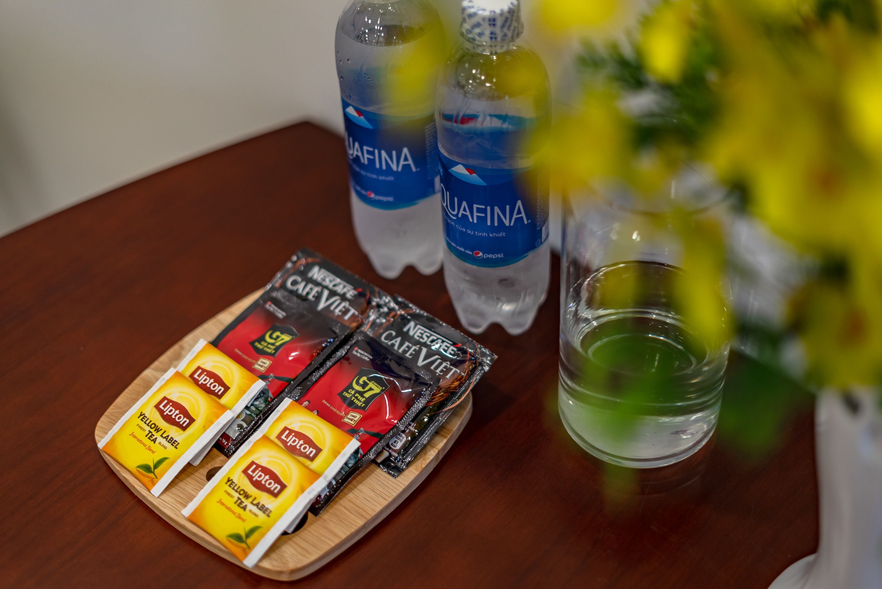 Una mesa de madera con una bandeja de sales de té Lipton, dos botellas de agua Aquafina, un vaso de líquido verde y una flor amarilla borrosa en primer plano.