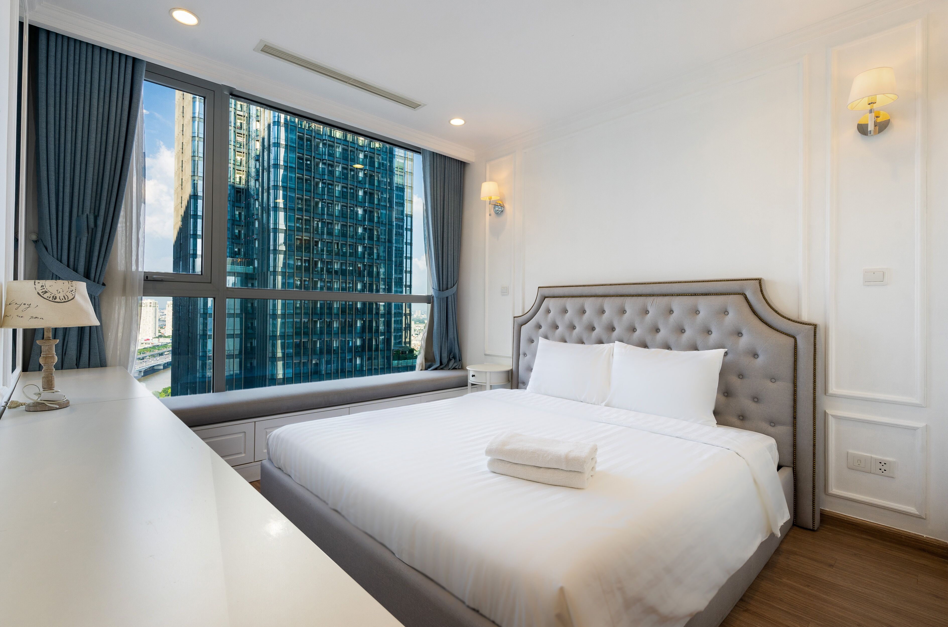 Habitación de hotel con una cama grande, una mesita de noche con lámpara, una ventana con cortinas y vistas a edificios altos.