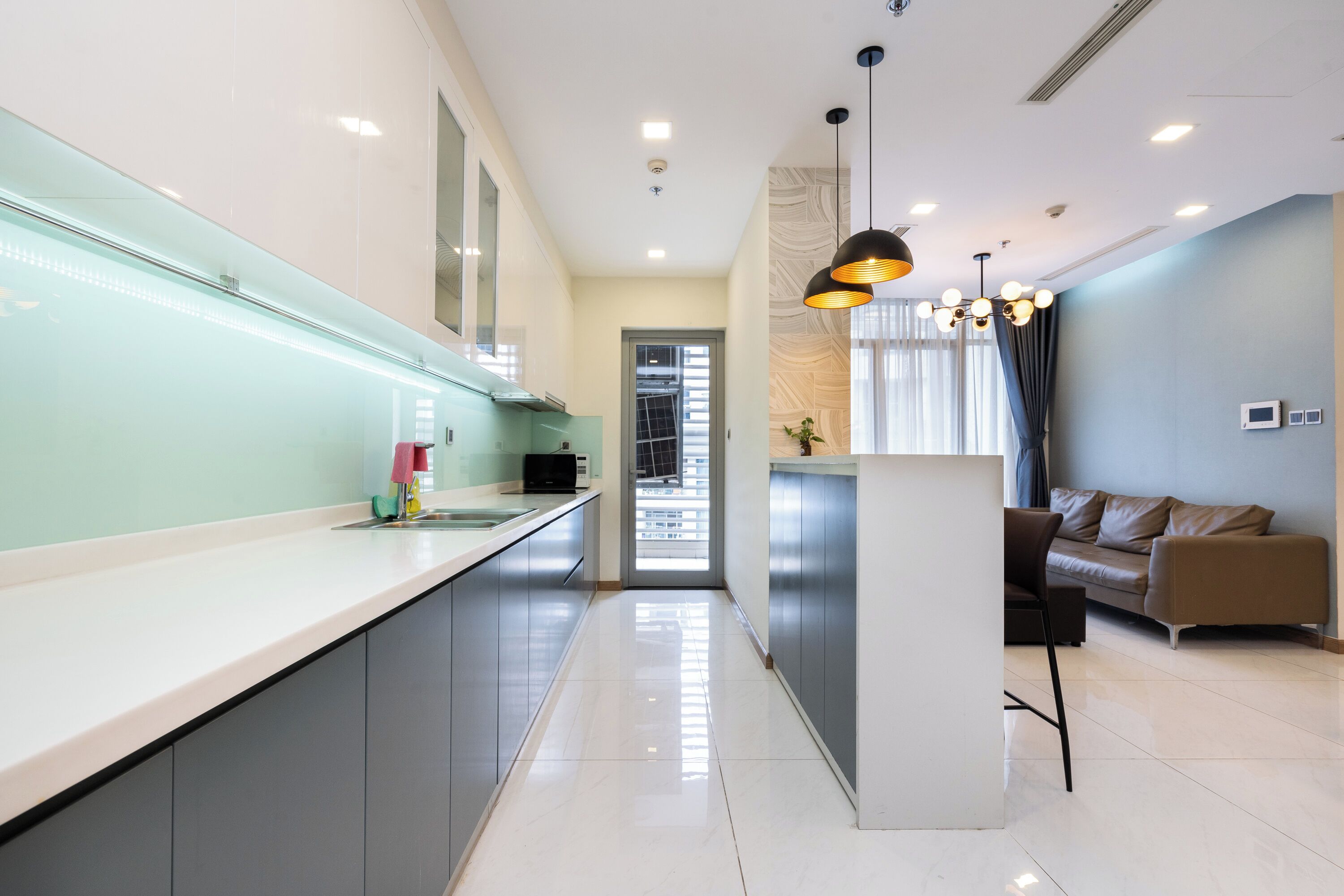 A modern kitchen with a long countertop, grey cabinets, and a central island. There is a dining area with a brown sofa and a pendant light.
