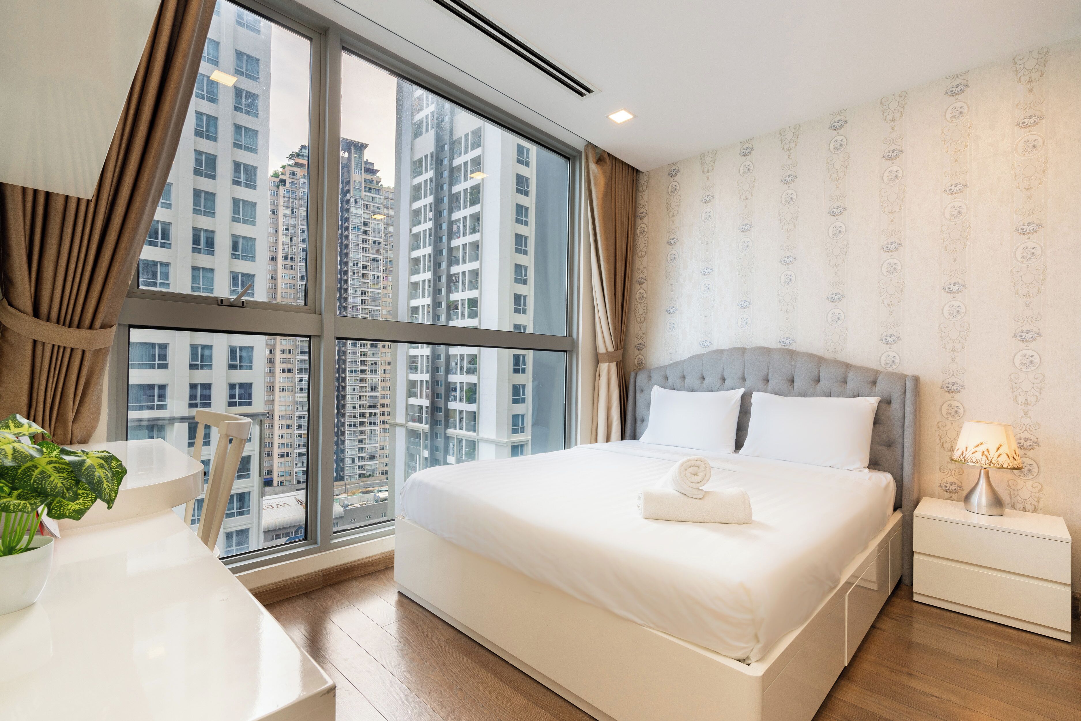 A modern hotel room with a large window, a bed with white linens, a bedside table with a lamp, and a view of high-rise buildings.
