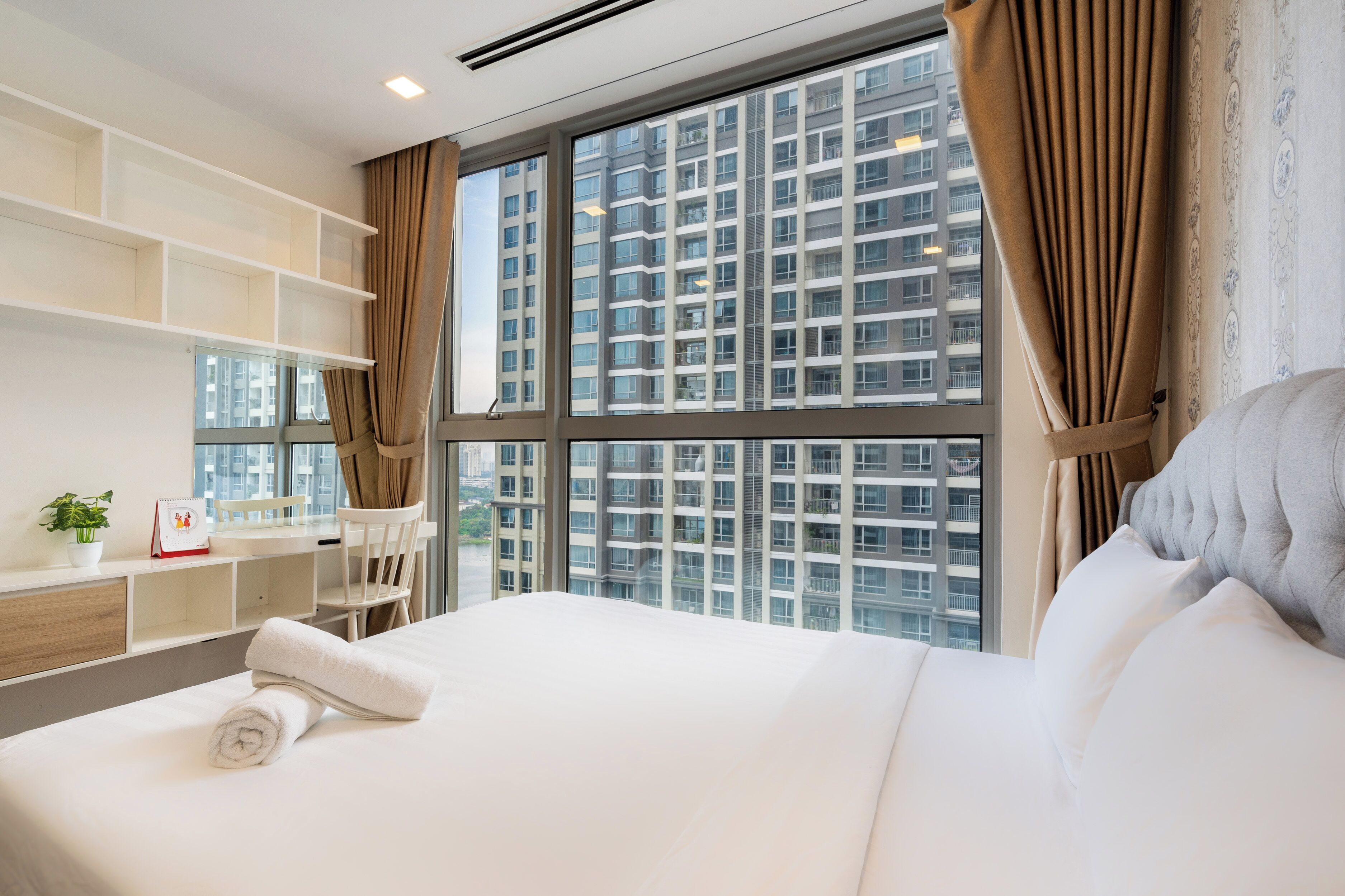 A modern hotel room with a large window, a neatly made bed, a small desk with a chair, and a shelving unit.
