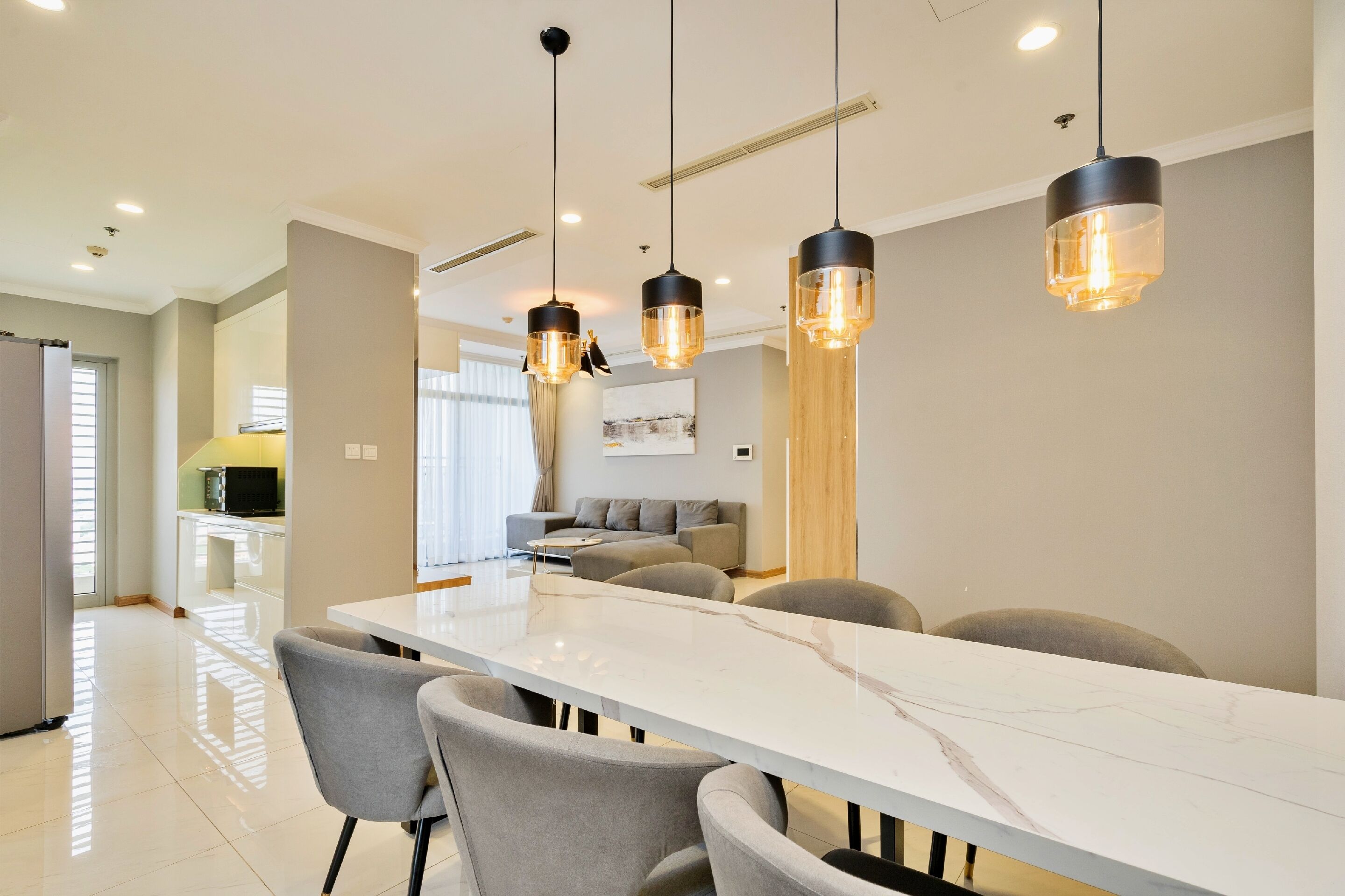 A modern dining area with a marble table and grey chairs, overhead pendant lighting, and a kitchen in the background.
