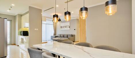 A modern dining area with a marble table and grey chairs, overhead pendant lighting, and a kitchen in the background.