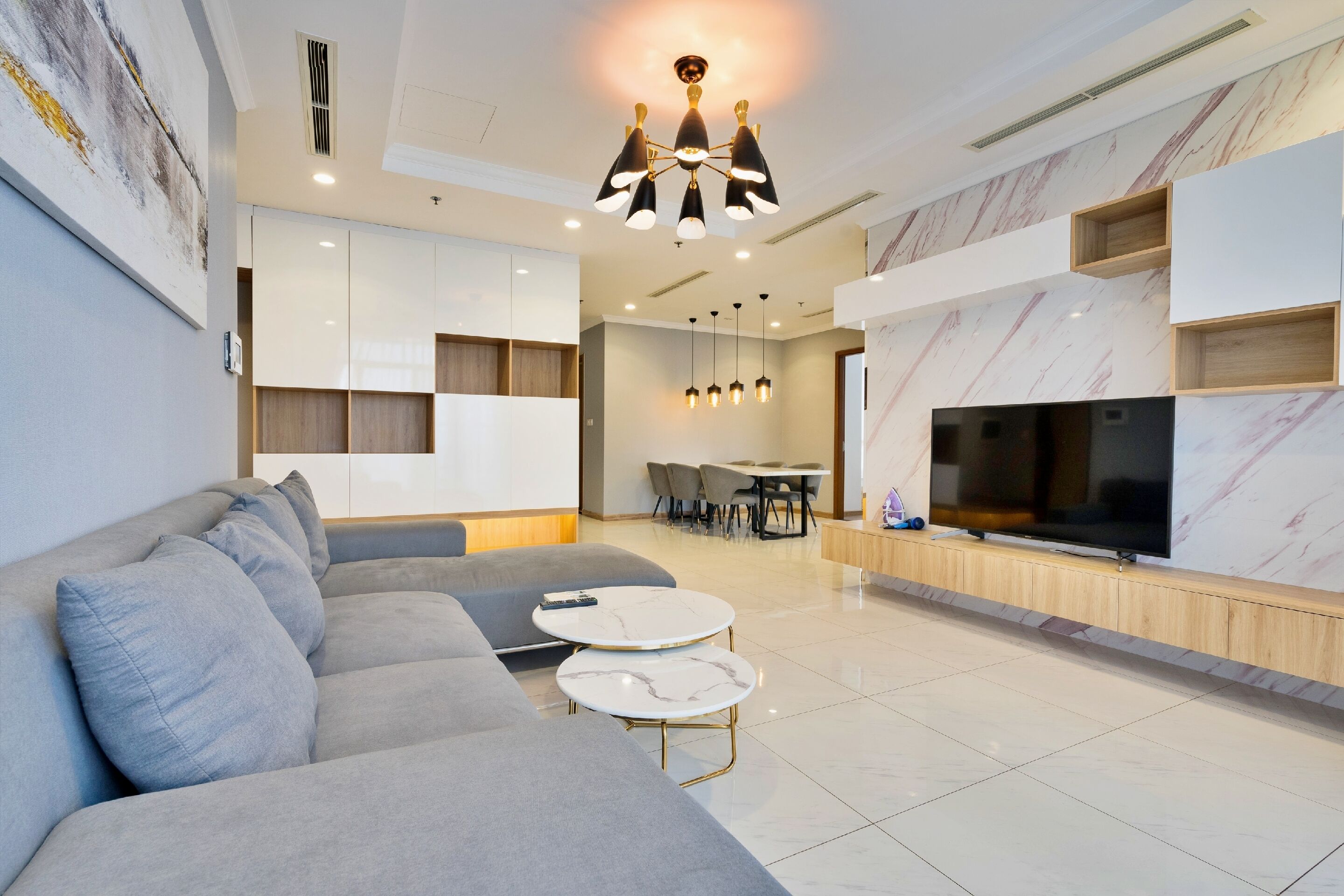 Una sala moderna con un sofá gris, dos mesitas de centro de mármol, un televisor de pantalla plana y un comedor con mesa y sillas.