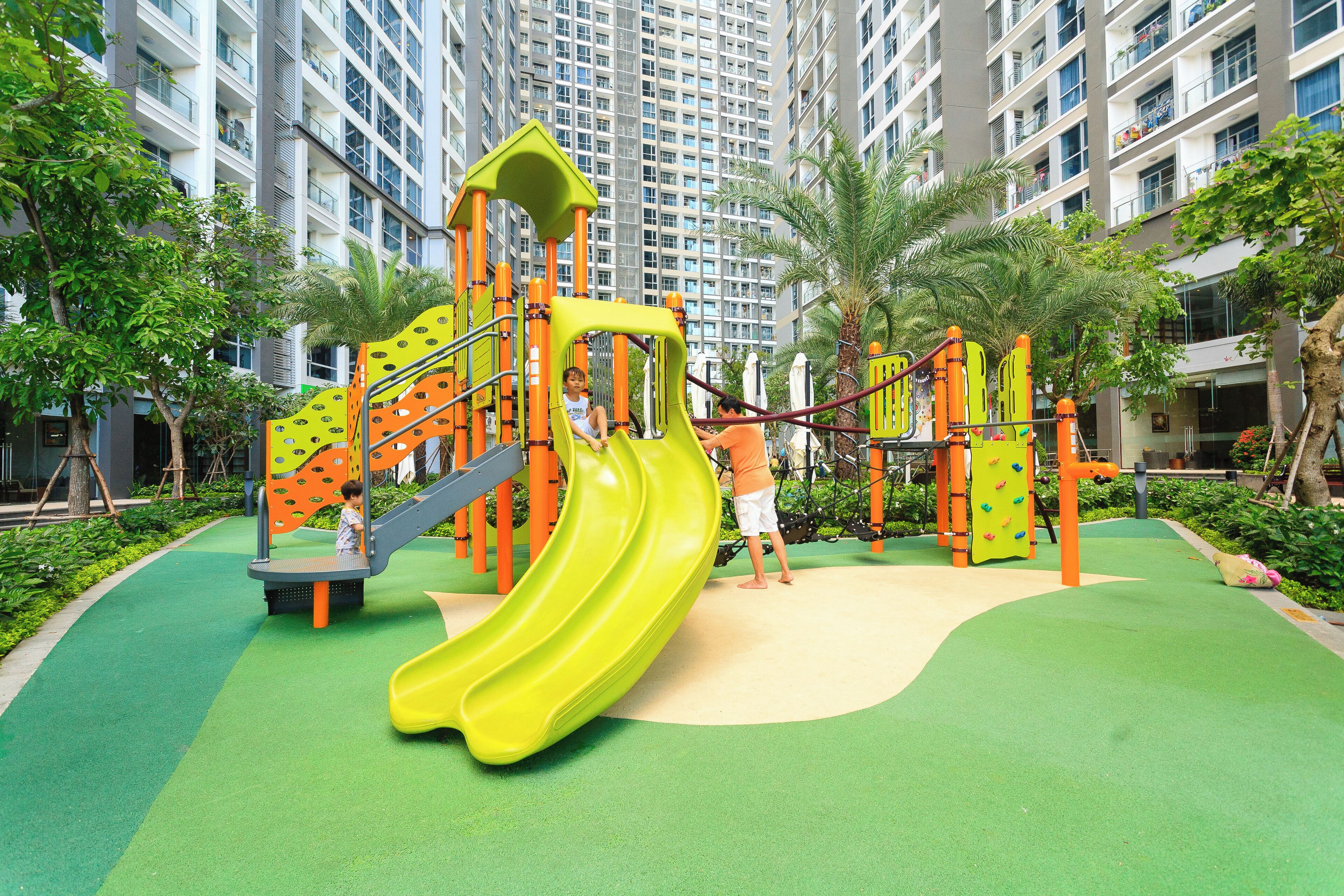 A playground with a yellow slide and climbing structures, surrounded by greenery and tall buildings.