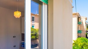 Ein modernes Hotelzimmer mit einem großen Schiebeglasfenster, das auf einen Balkon führt, einer Kristallleuchtre und Blick auf benachbarte Gebäude.