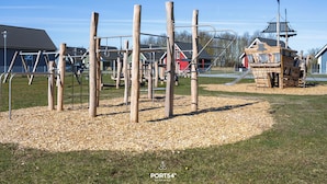Un parque infantil con estructuras de madera y un tobogán, rodeado de césped y acolchado de mantillo.