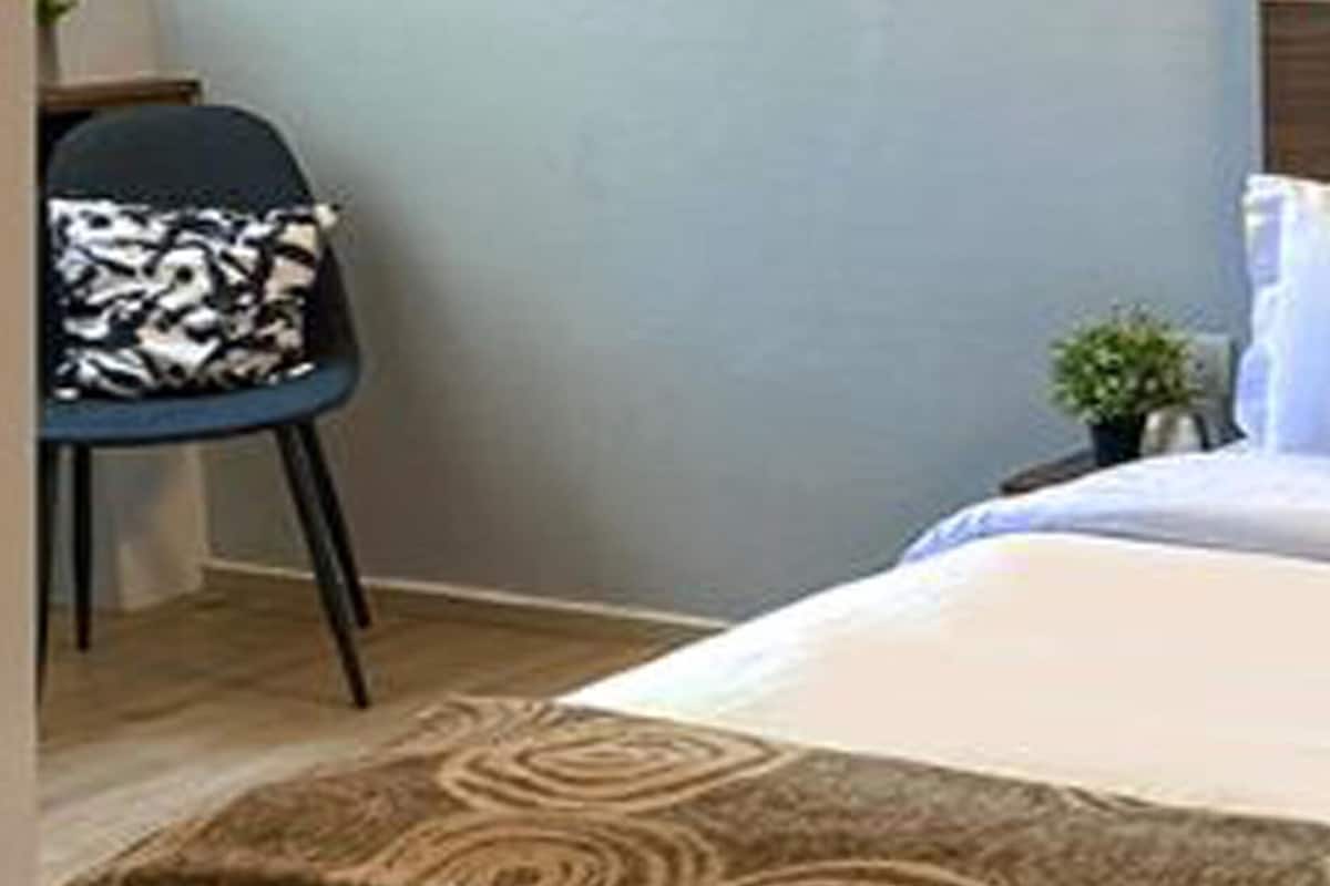 A neatly made bed with a patterned bedspread and decorative pillows, a small side table with a plant, and a chair with a patterned cushion in a room with light-colored walls and a wooden headboard.