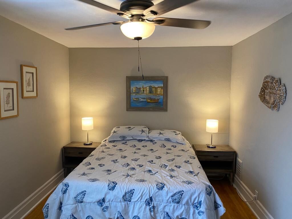 Une chambre à coucher comprenant un lit, deux tables de chevet, un ventilateur de plafond, un tableau au mur et deux lampes fixées au mur.
