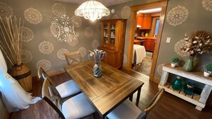 Un coin repas avec une table en bois, des chaises et un lustre. On peut apercevoir la cuisine à travers une porte ouverte.