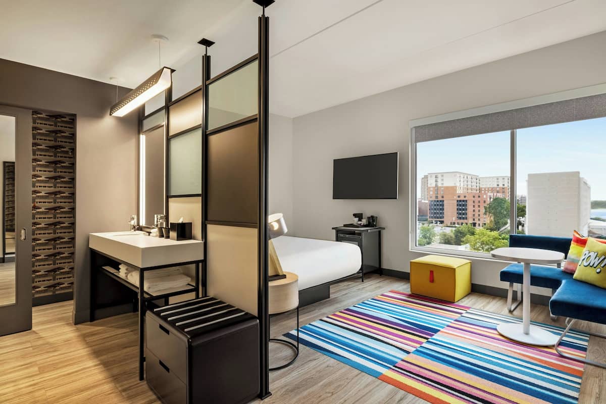 A modern hotel room with a large window, a bed, a desk, a chair, a TV, and a colorful striped rug.